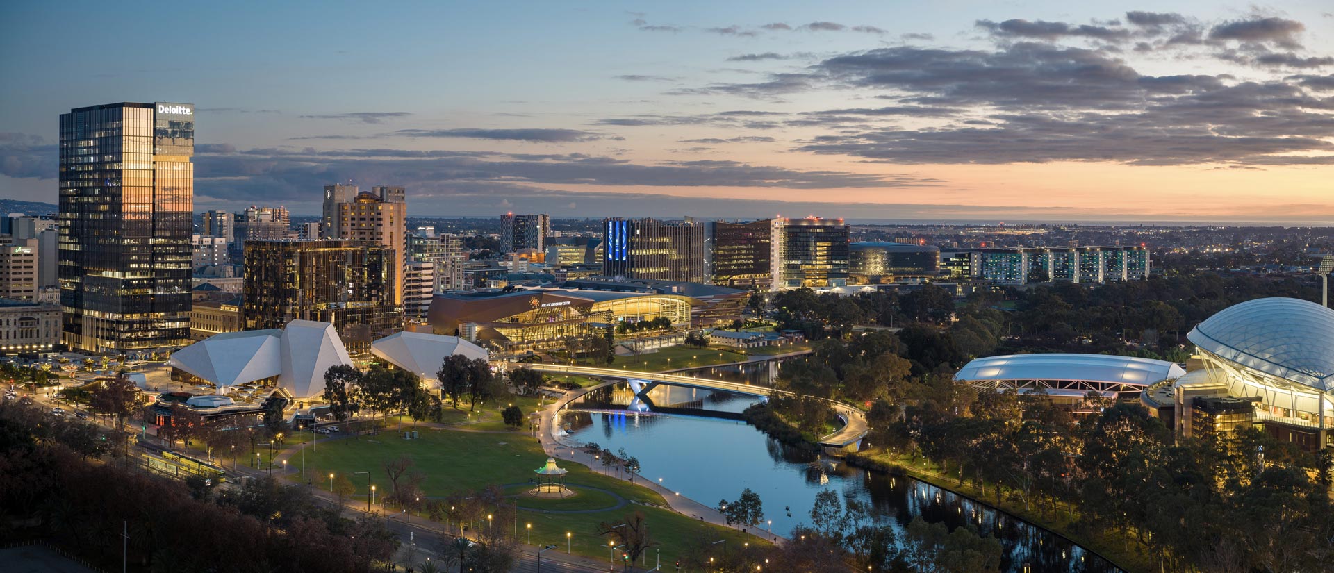 Adelaide CBD and riverfront at dusk, as viewed from a drone. Landmarks including Elder Park, the Convention Centre, Torrens Footbridge, Adelaide Oval and Royal Adelaide Hospital are visible with a partly cloudy sunset in the distance.
