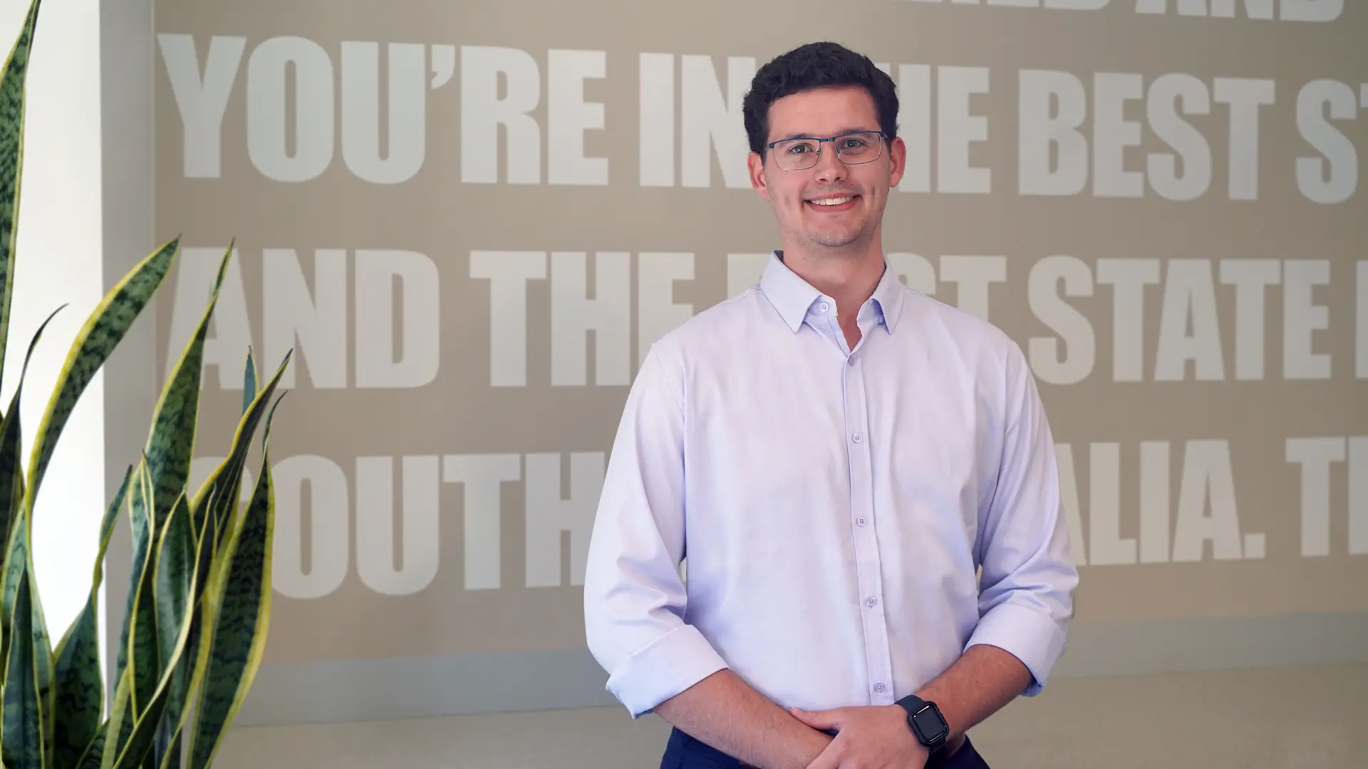Young man in light blue shirt stands in front of grey background and smiles at the camera
