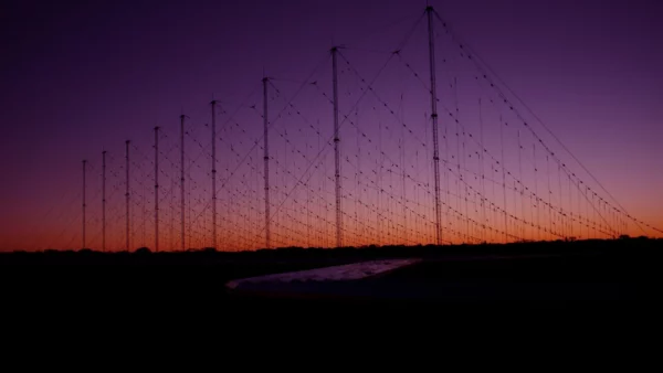 Silhouetted array of tall antenna towers with crisscrossing cables against a purple and orange sunset sky, with a dark foreground and low horizon line.
