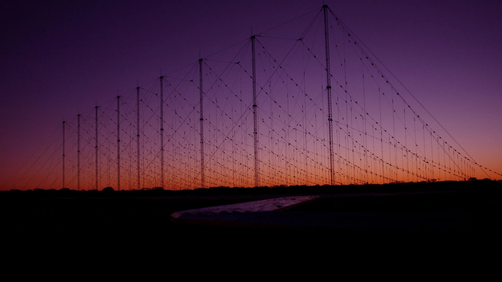 Silhouetted array of tall antenna towers with crisscrossing cables against a purple and orange sunset sky, with a dark foreground and low horizon line.