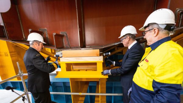 A man watches on as two others put a coin under the keel of a frigate ship under construction.