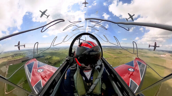 Pilot wearing a red flight helmet seated in the cockpit of an aerobatic aircraft, with five other aircraft flying in close formation overhead and alongside above a patchwork rural landscape under a partly cloudy sky.
