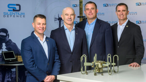 Four people in business attire stand behind a table displaying a camouflage-coloured radar device with multiple antenna loops at a Silentium Defence exhibition booth.